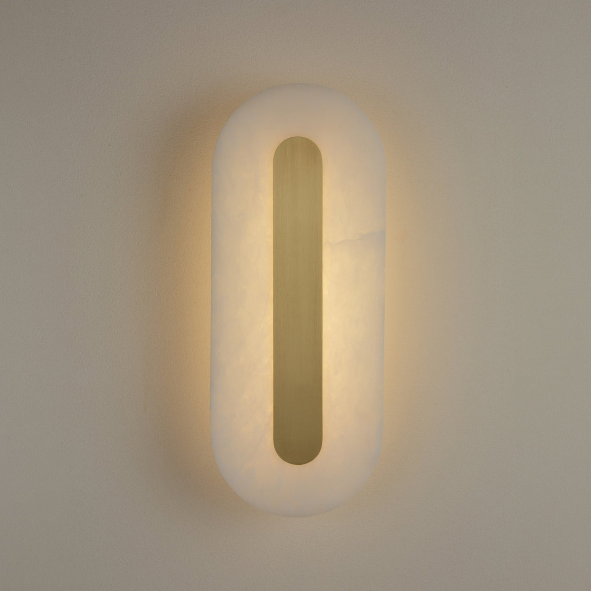 Averra medium alabaster wall light by Niori, illuminated pill-shaped panel with recessed brass spine casting warm white glow on a neutral wall.