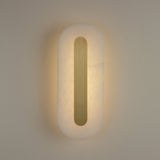 Averra medium alabaster wall light by Niori, illuminated pill-shaped panel with recessed brass spine casting warm white glow on a neutral wall.