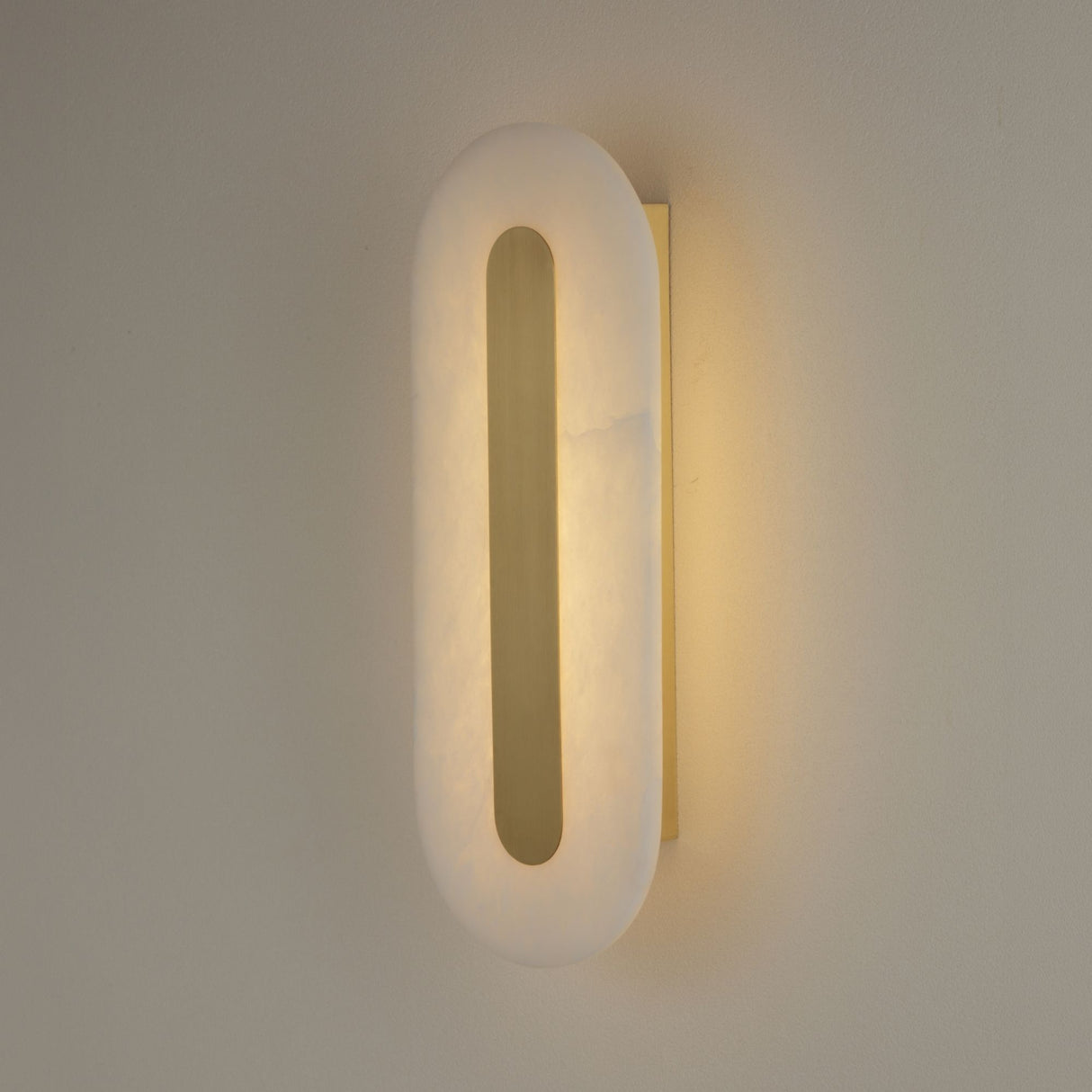 Averra LED large oval alabaster wall light from Niori, mounted on a neutral wall with a brushed brass spine glowing warm white through the stone.