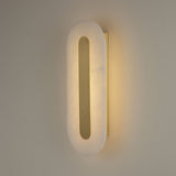 Niori Averra 800mm alabaster wall light illuminated on a pale wall, showing the elongated rounded soft white panel with a recessed brass linear centre.