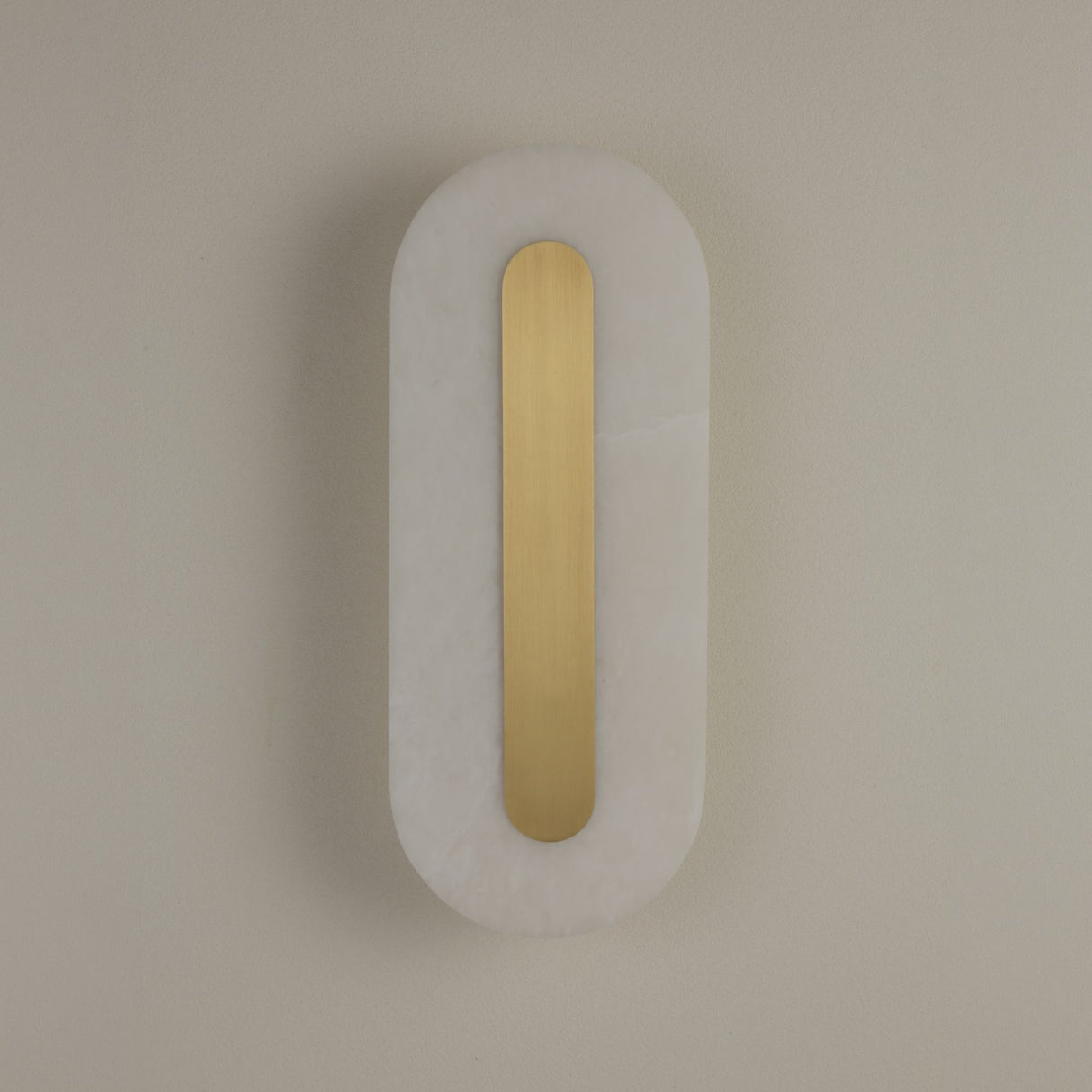 Niori Averra medium LED wall light with an elongated pill-shaped alabaster panel in soft white and a recessed brass spine, mounted on a neutral wall.