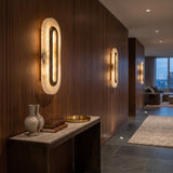 Ovaline LED Oval Alabaster Wall Light 46cm in Brushed Brass, glowing stadium-shaped alabaster plates with hammered brass rings lighting a wood-panelled hallway.