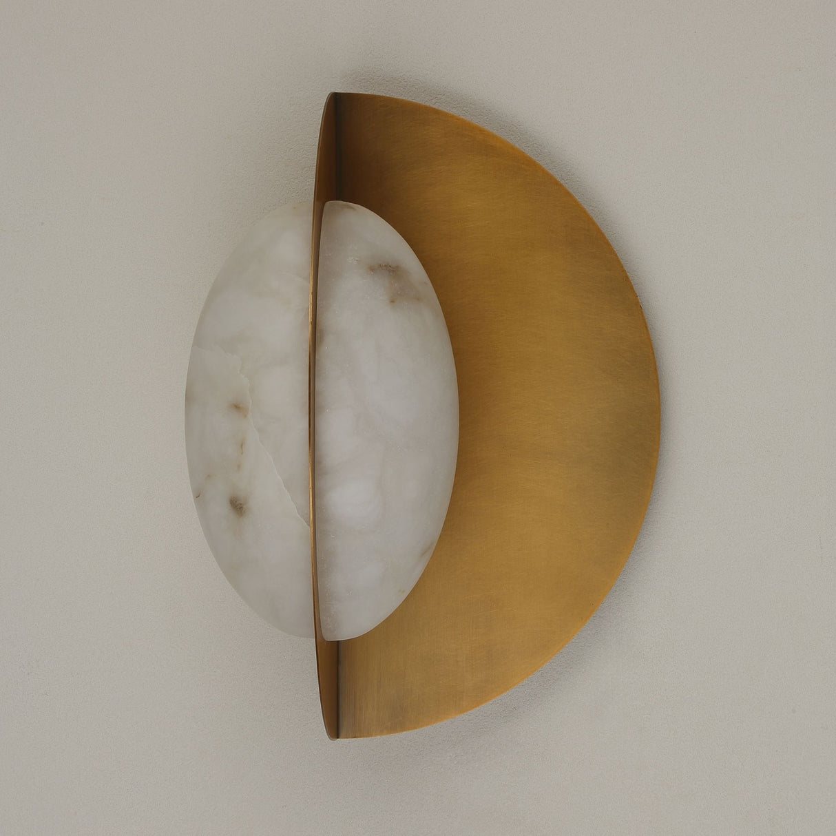 Niori Althia LED wall light with a rounded natural alabaster diffuser set against a brushed brass half-moon backplate, shown side-on against a pale wall.