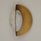 Niori Althia LED wall light with a rounded natural alabaster diffuser set against a brushed brass half-moon backplate, shown side-on against a pale wall.