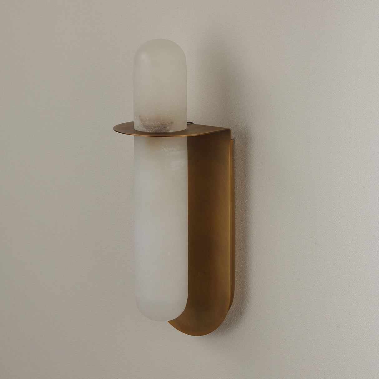 Niori Luneth Small LED wall light with a cylindrical natural Spanish alabaster column set in a curved brushed brass cradle, mounted on a pale wall.