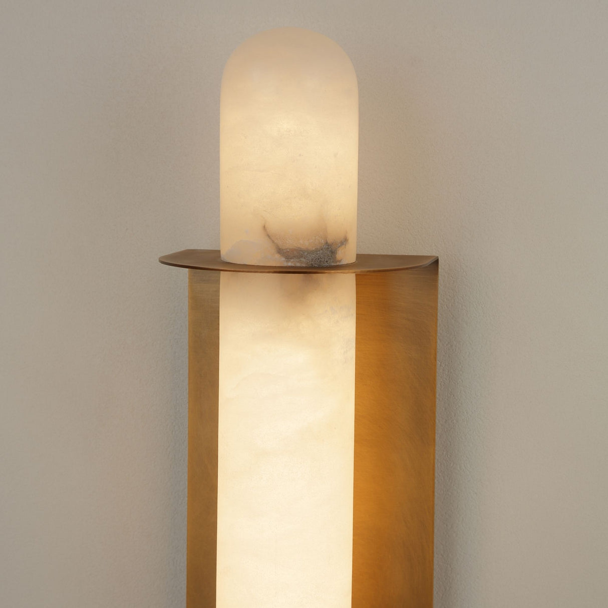 Luneth Small alabaster wall light by Niori with a rounded natural stone column set within a curved brushed brass cradle, glowing warm white.