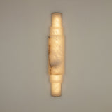 Niori Aurelia LED wall light in natural Spanish alabaster with stacked cylindrical sections and brushed brass end caps, illuminated against a neutral wall.