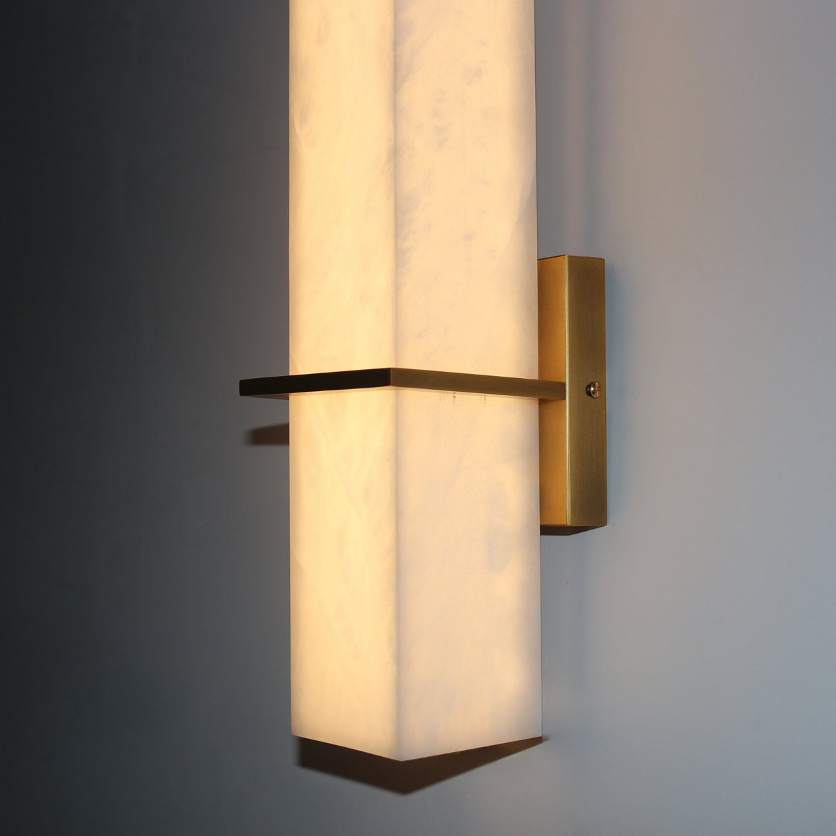 Aurevon LED Alabaster Vertical Panel Wall Light in Brushed Brass, close-up of the illuminated stone panel and horizontal brass arm against a grey wall.