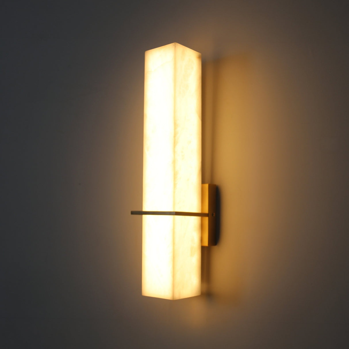 Aurevon LED alabaster vertical panel wall light with brushed brass backplate and slim horizontal arm, glowing warm white against a dark background.