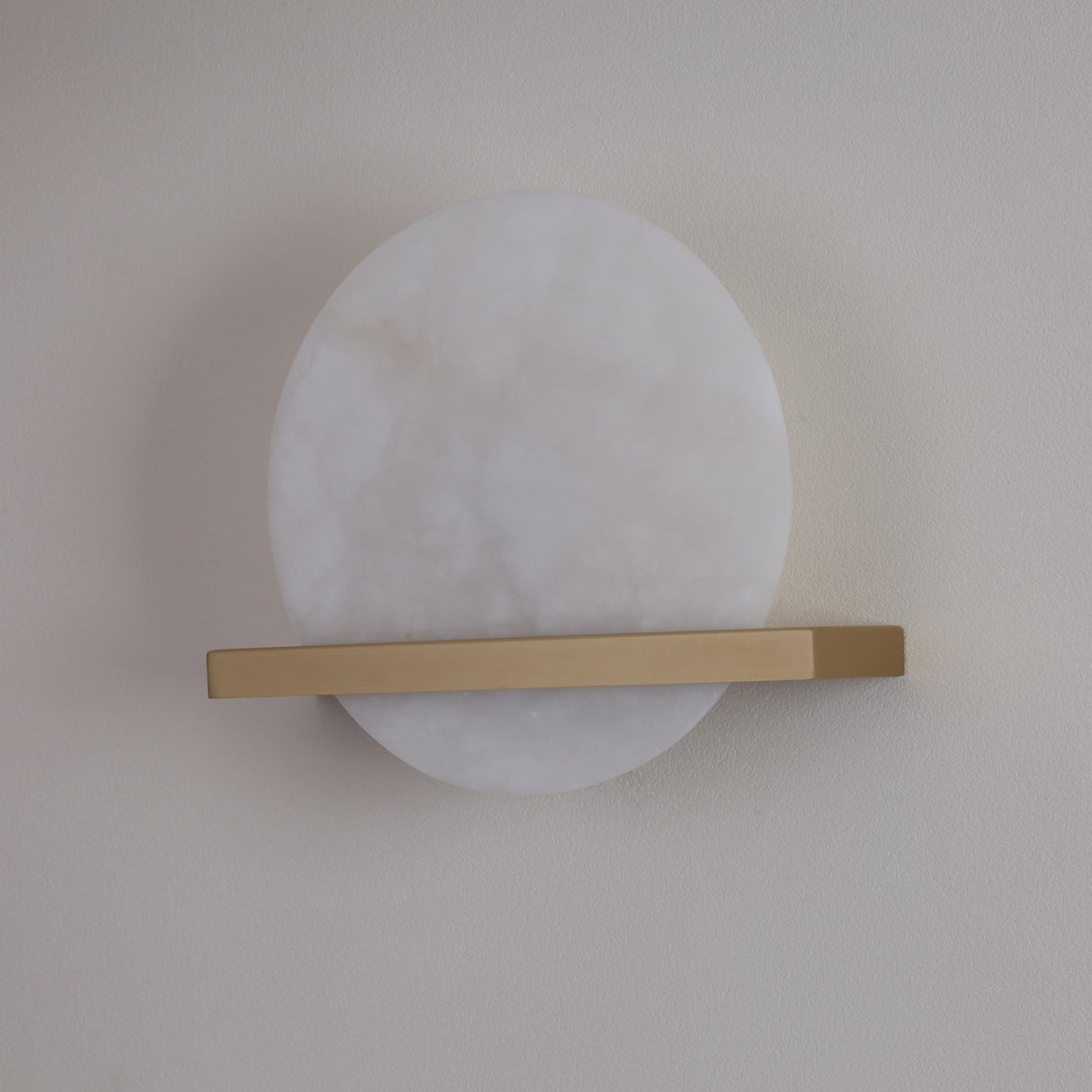 Niori Caldris LED circular alabaster wall light with a white stone disc bisected by a slim brushed brass bar, mounted on a pale wall.