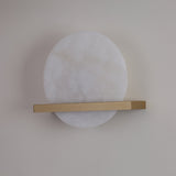 Niori Caldris LED circular alabaster wall light with a white stone disc bisected by a slim brushed brass bar, mounted on a pale wall.