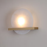 Niori Caldris circular alabaster wall light illuminated with warm LED glow, bisected by a brushed brass bar, mounted on a neutral wall.