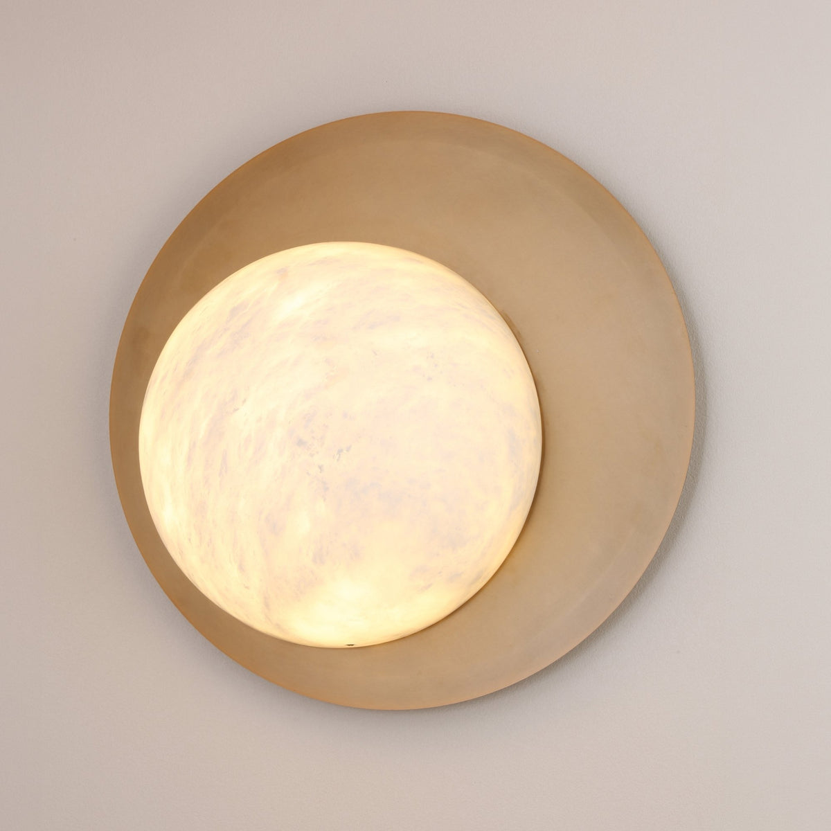 Niori Lunara round wall light with hand-cut Spanish alabaster disc on a brushed brass backplate, glowing warmly against a light grey wall.