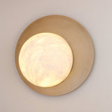 Niori Lunara round wall light with hand-cut Spanish alabaster disc on a brushed brass backplate, glowing warmly against a light grey wall.