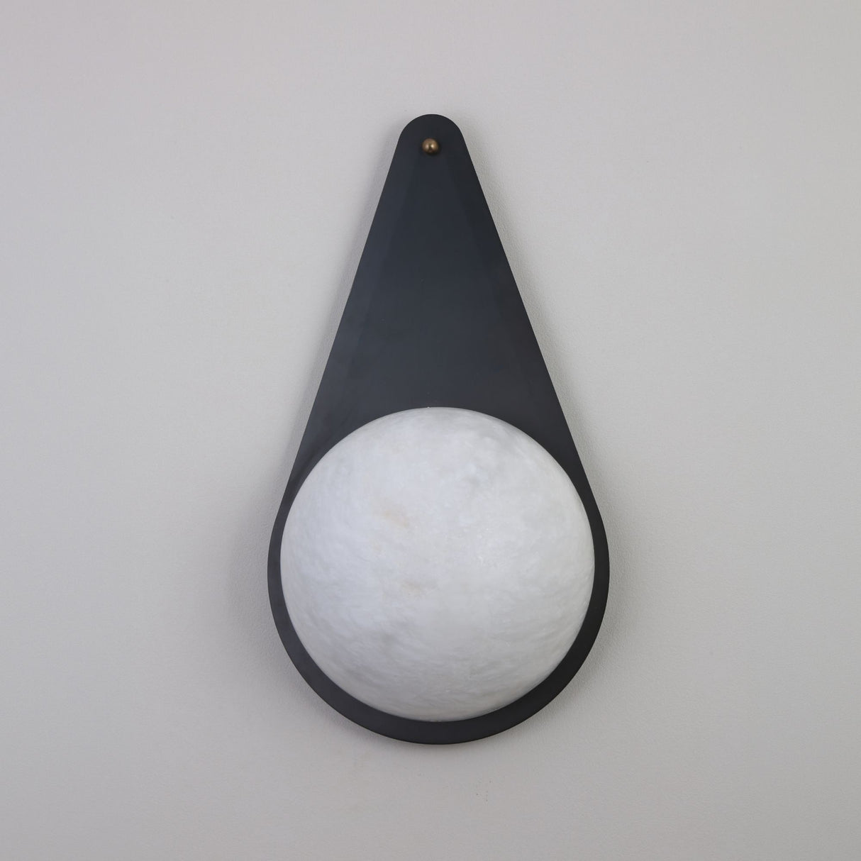 Niori Lunara teardrop wall light with a matt black copper backplate, brass pin accent, and natural Spanish alabaster disc, shown on a light grey wall.