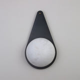 Niori Lunara teardrop wall light with a matt black copper backplate, brass pin accent, and natural Spanish alabaster disc, shown on a light grey wall.