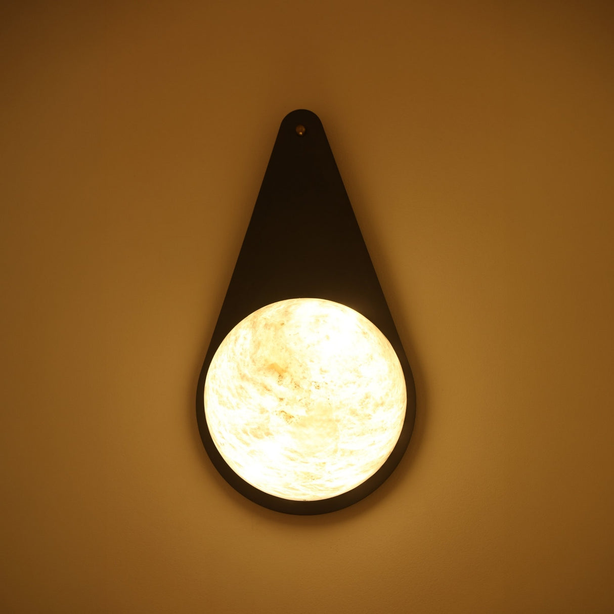 Niori Lunara teardrop wall light with matt black backplate and illuminated natural alabaster disc showing cream veining, mounted on a warm-toned wall.