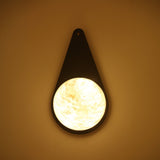 Niori Lunara teardrop wall light with matt black backplate and illuminated natural alabaster disc showing cream veining, mounted on a warm-toned wall.