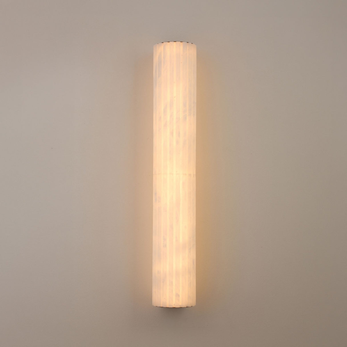 Niori Zenvia LED wall light illuminated, showing a tall vertical alabaster column with soft warm glow and subtle veining, mounted on a light grey wall.