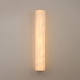 Niori Zenvia LED wall light illuminated, showing a tall vertical alabaster column with soft warm glow and subtle veining, mounted on a light grey wall.