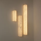 Niori Aureline LED alabaster column wall lights grouped at varying heights on a pale corner wall, glowing warm white through natural Spanish stone.