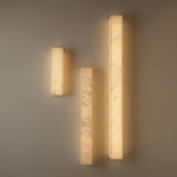 Three Niori Aureline alabaster column wall lights in varying heights, glowing warm white against a pale grey wall.