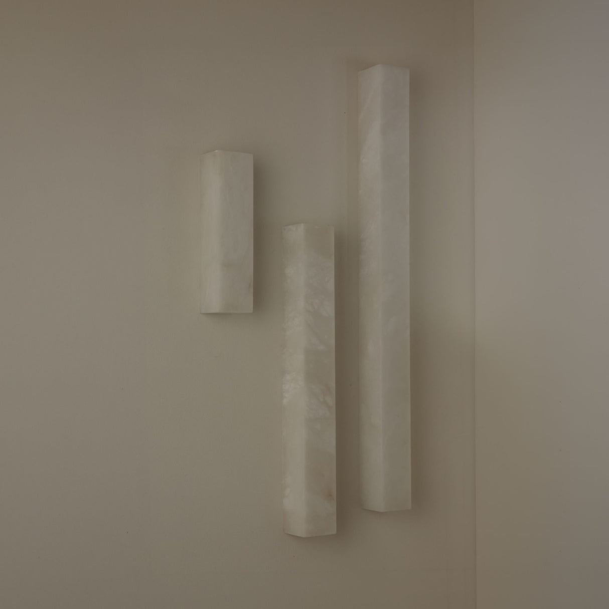 Three Niori Aureline alabaster column wall lights in varying heights with soft white stone and brass edges, mounted on a neutral corner wall.