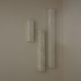 Three Niori Aureline alabaster column wall lights in varying heights with soft white stone and brass edges, mounted on a neutral corner wall.