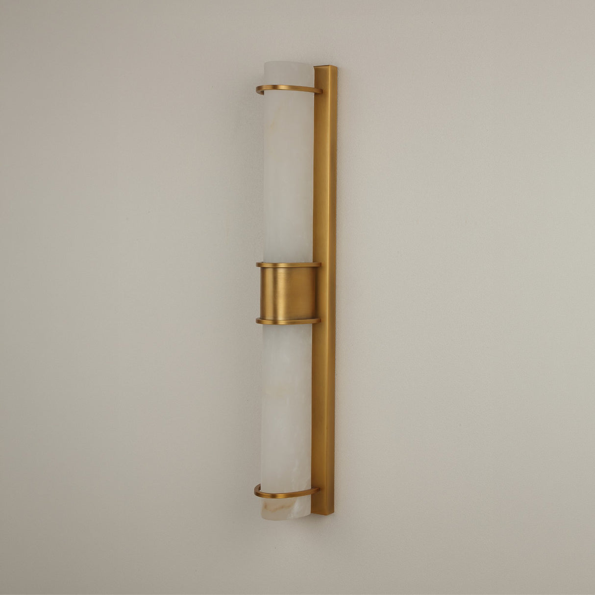 Niori Averna LED framed wall light with two stacked natural alabaster cylinders held in a brushed brass frame and central collar, mounted on a pale wall.