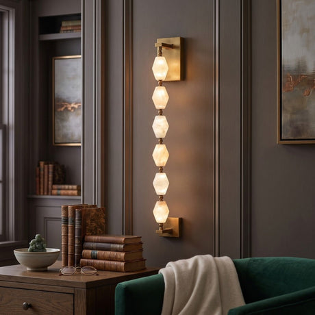 Brisalda 6 Light Faceted Alabaster Wall Light in Brushed Brass, vertical hexagonal stone column mounted on a study wall beside books and a green armchair.