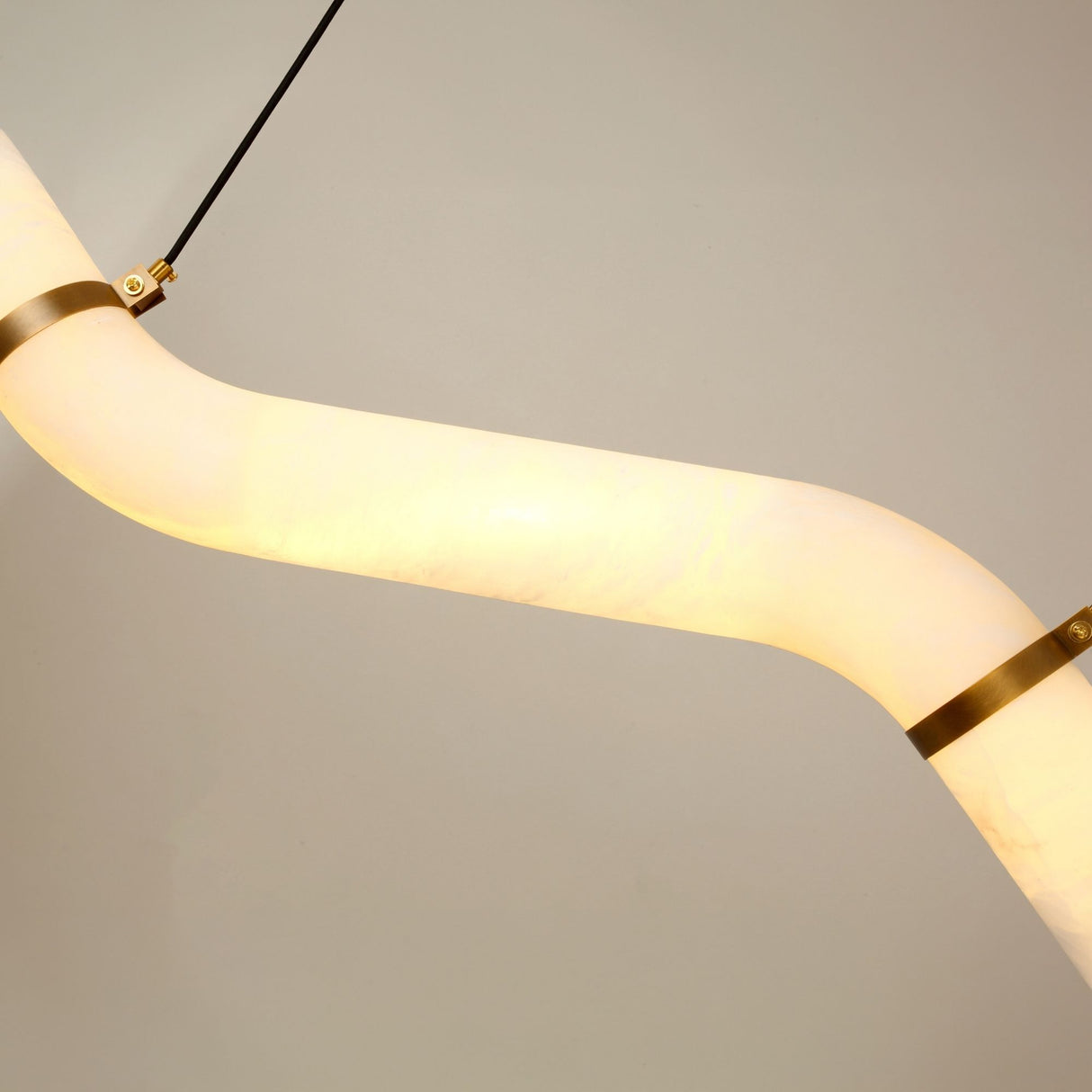Close-up of the Zareon Small LED chandelier's stepped alabaster tube glowing warm white, joined by brushed brass collars and black cable drop, against a plain grey background.