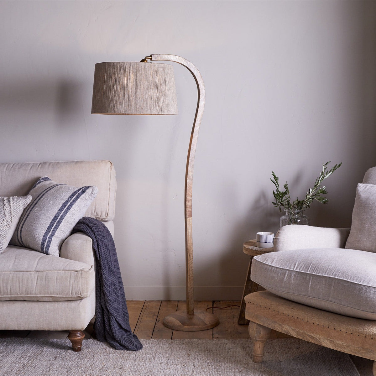 Namira Mango Wood Floor Lamp with curved arching post, natural wood finish, and fabric drum shade, shown beside a beige sofa in a living room setting.
