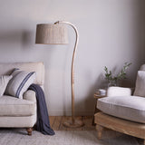 Namira Mango Wood Floor Lamp with curved arching post, natural wood finish, and fabric drum shade, shown beside a beige sofa in a living room setting.