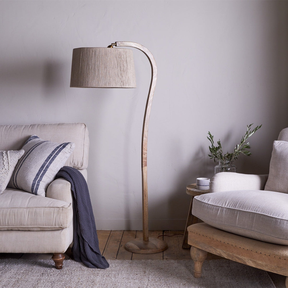 Namira Mango Wood Floor Lamp Base in natural wood with an arched post and fabric drum shade, shown beside a beige sofa in a living room.