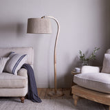 Namira Mango Wood Floor Lamp Base in natural wood with an arched post and fabric drum shade, shown beside a beige sofa in a living room.