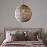 Nilaveli Woven Jute Sphere Pendant Light in natural jute on a single cord, shown hanging above a bed with brown cushions and neutral bedding in a bedroom setting.