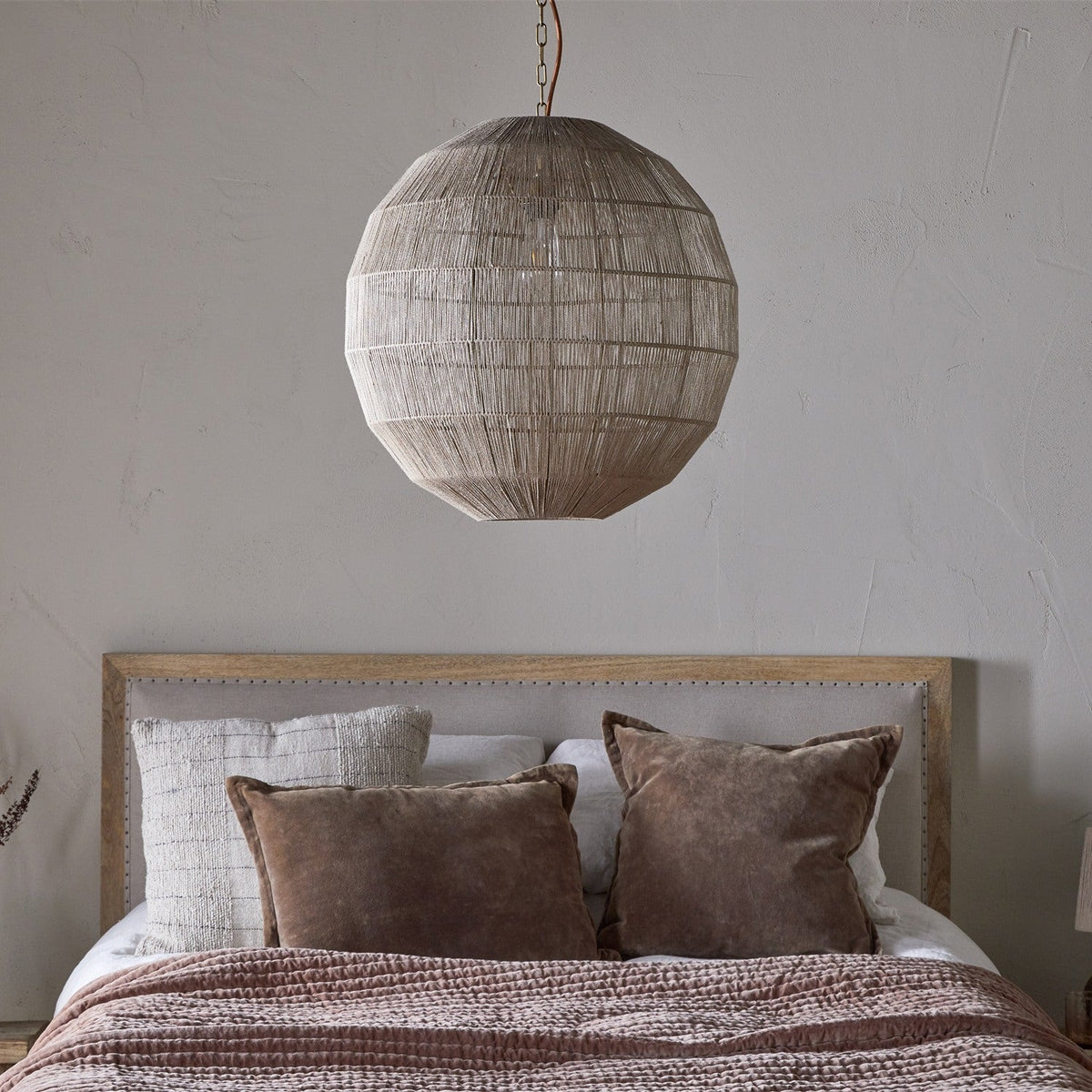 Nilaveli Woven Jute Sphere Pendant Light in natural jute hangs above a bed with brown velvet cushions and woven throw, set against a plain grey wall.