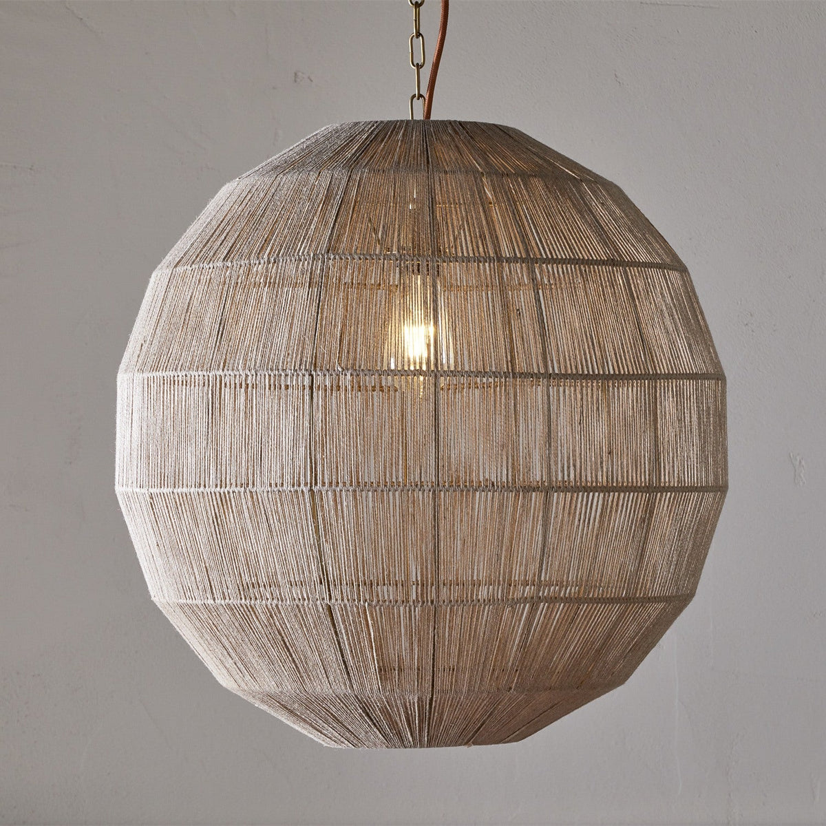Nilaveli Woven Jute Sphere Pendant Light in natural jute, spherical globe shade with visible ribbed frame lines, hung on a chain with a single exposed bulb, shown against a plain grey wall.