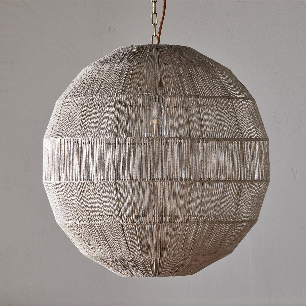 Nilaveli Woven Jute Sphere Pendant Light in natural tone, a single-bulb spherical shade wound in dense vertical jute threads with horizontal banding, hung from a chain against a plain grey wall.