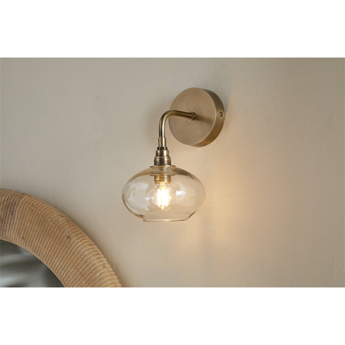 Otoro Recycled Glass Wall Light in Clear, shown on a neutral wall with brass round plate, curved arm, ribbed collar and flattened clear glass shade exposing a lit bulb.