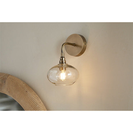 Otoro Recycled Glass Wall Light in Clear, shown on a neutral wall with brass round plate, curved arm, ribbed collar and flattened clear glass shade exposing a lit bulb.
