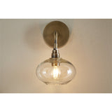 Otoro wall light with round brass mounting plate, curved arm, and flattened clear recycled glass shade on a ribbed brass collar, lit bulb visible, mounted on a plain wall.