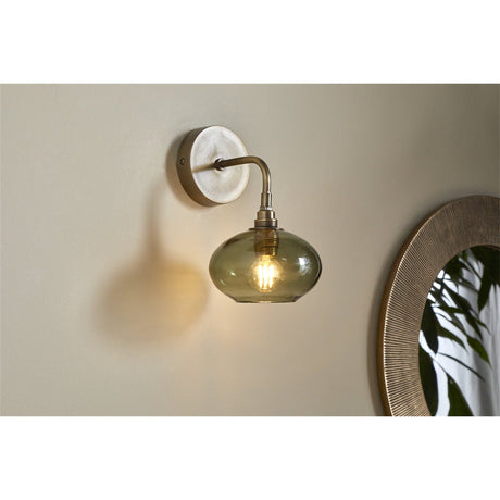 Otoro recycled glass wall light in green, with a round brass wall plate, curved brass arm, and flattened olive-green glass shade lit by a visible bulb, shown mounted beside a mirror.