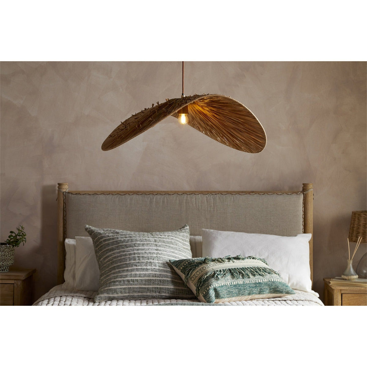 Essubi Raffia Organic Weave Pendant Light in Natural, a leaf-shaped folded raffia shade with a single E27 bulb, hung above a bed with upholstered headboard and cushions.