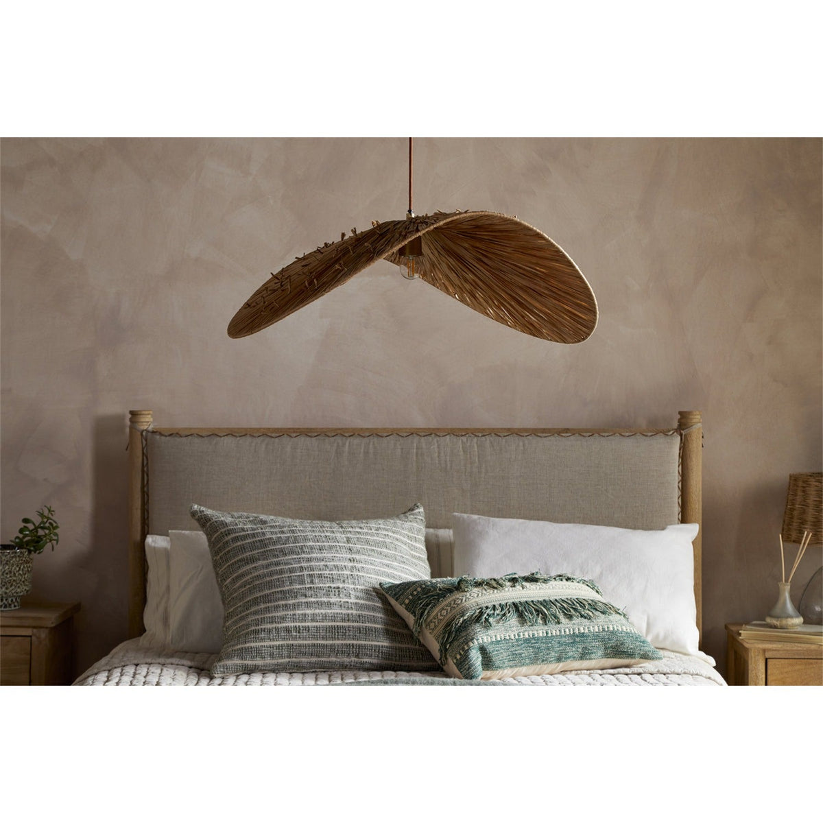 Essubi Raffia Organic Weave Pendant Light in natural raffia, a single folded leaf-shaped shade with visible rib strands, hung above a bed in a neutral-toned bedroom.
