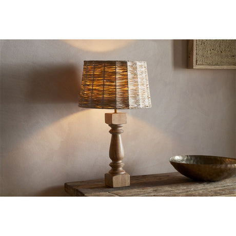 Hega Mango Wood Lamp Base in Natural with a turned baluster shaft and squared foot, shown lit with a woven tapered shade on a rustic wooden console.