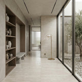 A modern, minimalist hallway features light stone floors, built-in shelves with pottery, a gray bench, the Bubble LED Glass Floor Lamp - Gold, and large windows overlooking a rock garden with an olive tree.