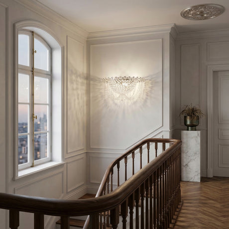 Elegant hallway with a wooden staircase, white panelled walls, large city-view window, and the Amara 2 Light K9 Crystal Flush Ceiling Light in chrome casting luminous patterns over a marble pedestal topped with a decorative vase of dried plants.