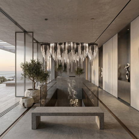 A modern, minimalist interior with a concrete bench, glass railings, the Symphonia 10 Light Metal Chain Veil Suspension in nickel, indoor trees, sculptures, and floor-to-ceiling windows overlooking the ocean and landscape.