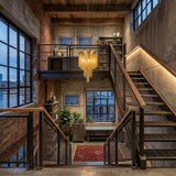 An industrial-style staircase with metal railings, exposed brick walls, large windows, and the Valora 5 Light Metal Chain Veil Chandelier in Gold. Plants, art, bookshelves, and a red rug complete the cozy urban loft vibe.